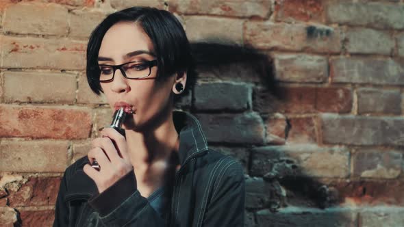 Gothic Woman with Short Black Hair Smokes an Electronic Cigarette on a Background of a Brick Wall