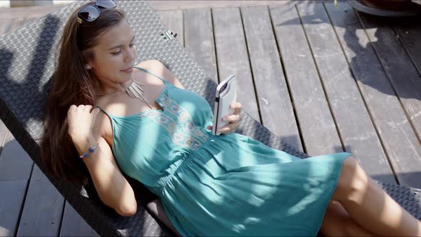 Woman in Blue Dress Resting in Lounge Chair and Using Smartphone