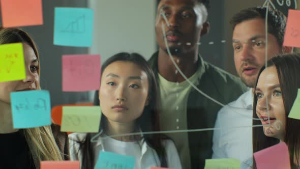 Diverse Coworkers Group Analyzing Corporate Strategy Drawing Chart on Glass Wall with Sticky Notes