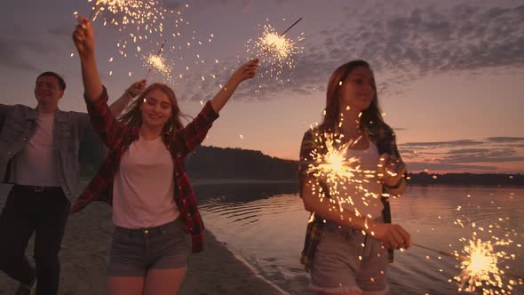 Cheerful Male and Female Friends are Running Along the Beach at Sunset Holding Sparkling Fireworks