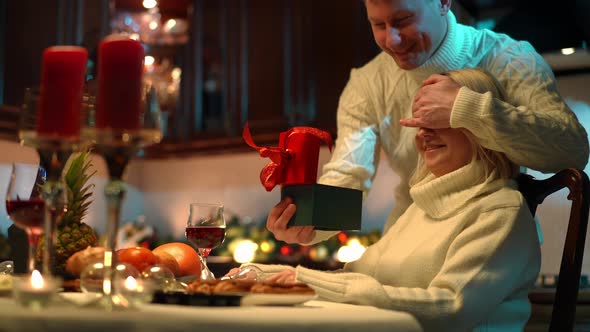 Smiling Woman Sitting on the Right As Man Covering Eyes with Hands Surprising Spouse with Gift