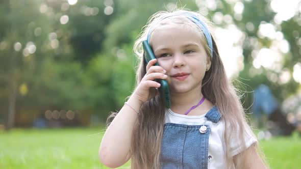 Happy little child girl having conversation talking on her sellphone in summer park. alt
