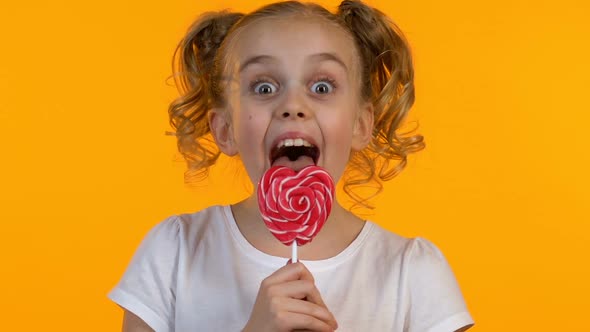 Cute Girl With Pigtails Licking Heart-Shaped Candy, Isolated Yellow Background