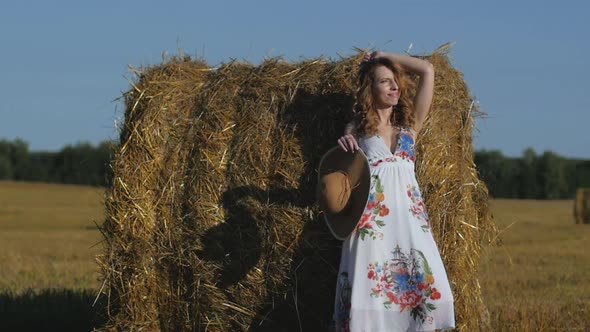 Beautiful Redhead Woman in Dress and Hat Stands Near a Haystack