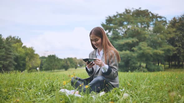 A Young Girl Makes a Purchase with Her Phone and Bank Card in the Park Sitting on the Grass