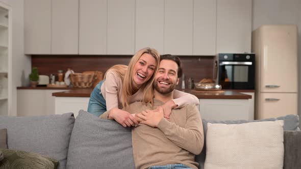 Cheerful Caucasian Couple in Love Sitting on the Sofa in Embrace and Looking at the Camera