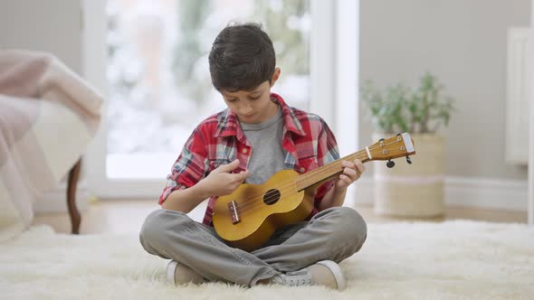 Happy Relaxed Middle Eastern Boy Playing Ukulele Sitting on Carpet at Home