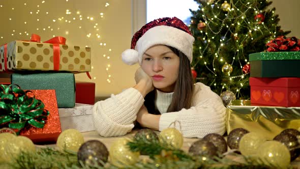 Sad Woman in a Santa Hat Sits Alone Against the Background of a Christmas Tree and Boxes with Gifts