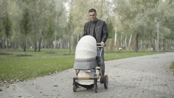 Portrait of Young Handsome Caucasian Man Walking with Baby Stroller Along Park Alley