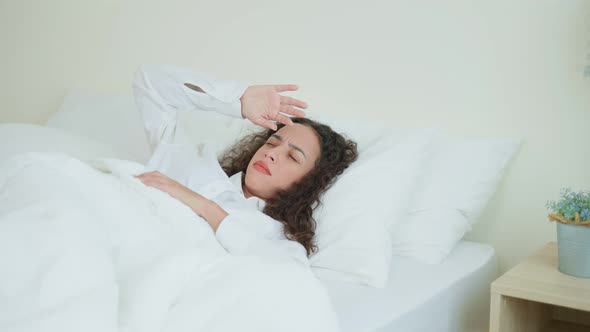 Latino beautiful girl in pajamas feel sick while sit on bed in bedroom.