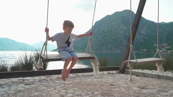 A Boy Swings on a Swing Near the Sea with a Beautiful View of the Bay and Mountains