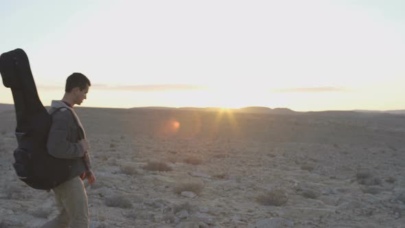 Young man walks with guitar case in desert sunset