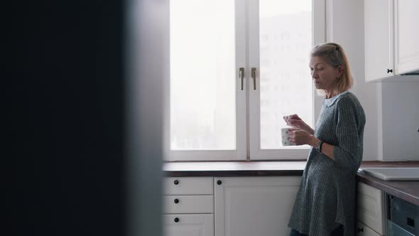 Depressed Lonely Elderly Woman Mixing Sugar in the Hot Beverage While Standing in Her Kitchen