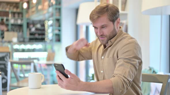 Young Man Celebrating Success on Smartphone in Cafe