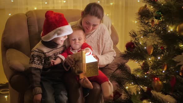 Happy Family with Children Sitting in Armchair at Christmas Tree and Looking Inside Glowing