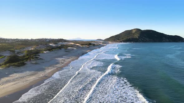 Aerial drone view flying over the beach with people walking in the late afternoon with waves, dunes