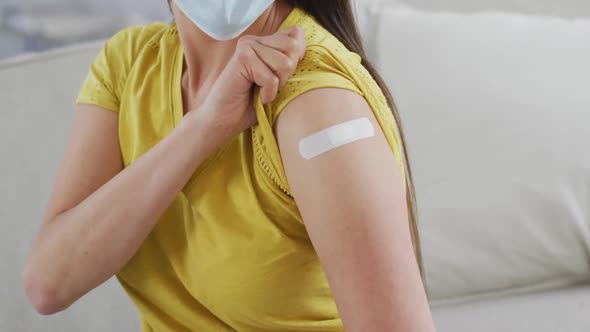 Midsection of asian woman in face mask sitting on sofa showing arm with plaster after vaccination