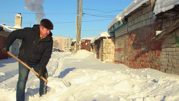 A Man in Warm Clothes with a Shovel Clears the Road of the Snow