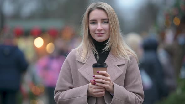 Portrait of a young girl in a coat standing on street and holding cup of hot drink.