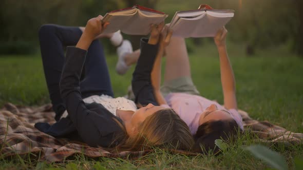 Curios Absorbed Teenage Girls Reading Books Lying on Blanket on Green Summer Spring Lawn at Golden