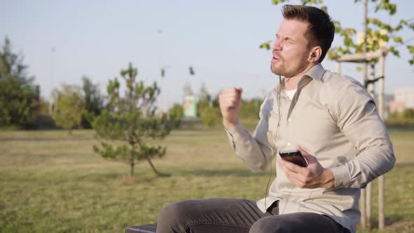 A Young Caucasian Man Listens to Energetic Music with Earphones on a Smartphone As He Sits in a Park