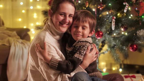 Portrait of Cheerful Smiling Boy with Mother Hugging Next to Christmas Tree and Looking in Camera