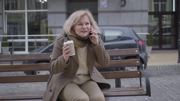 Portrait of Excited Senior Caucasian Woman Talking on the Phone As Sitting on Bench Outdoors
