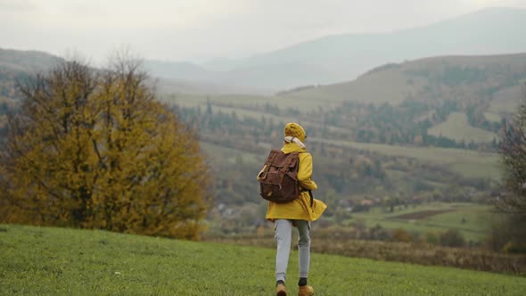 Slow Motion Happy Woman Hiker in Yellow Raincoat with Backpack Runs Away with Raised Arms in Green