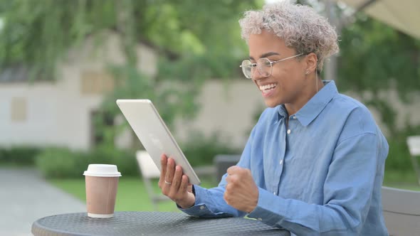 Successful Young African Woman Celebrating on Tablet in Outdoor Cafe