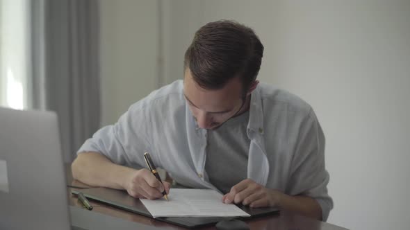 Man Sitting at the Table Writing Something on the Paper at Home