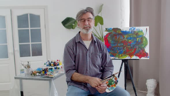 Male paint artist with beard is sitting on chair in studio and smiling, holding brushes and palette