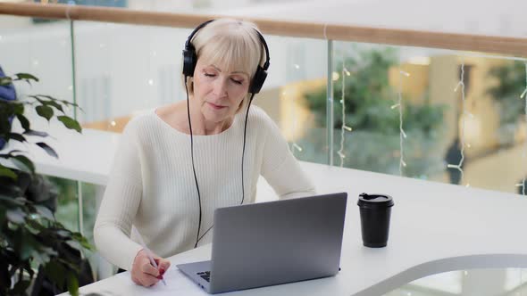 Mature Caucasian Woman Businesswoman Teacher in Headphones Looks at Laptop Screen Takes Notes Writes