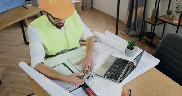 Designer in Helmet and Vest which Working with Architectural Scheme on Blueprint in Workroom