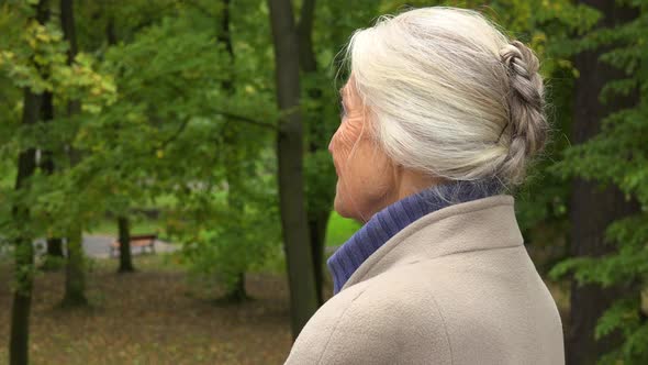 An Elderly Woman Looks Around Thoughtfully in a Park Closeup