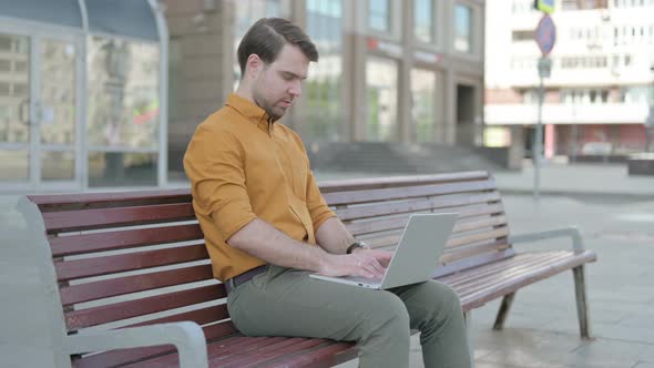 Busy Young Man Using Laptop Sitting Outdoor on Bench