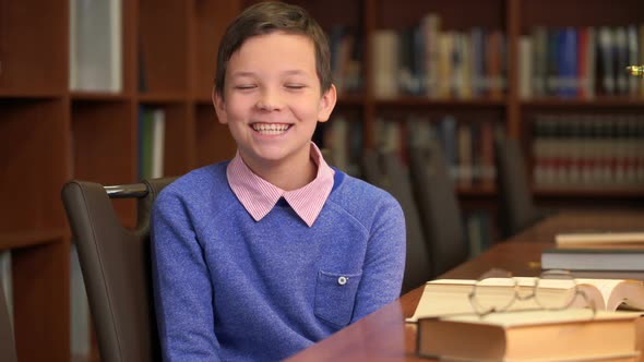 Portrait Shot of the Cute Schoolboy Sits Near the Bookshelf in the Library