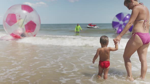 The Kid Looks at the Horizon of the Sea and Inflatable Big Balls with His Mother in a Bikini