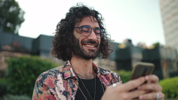 Smiling curly-haired bearded man in eyeglasses texting by mobile