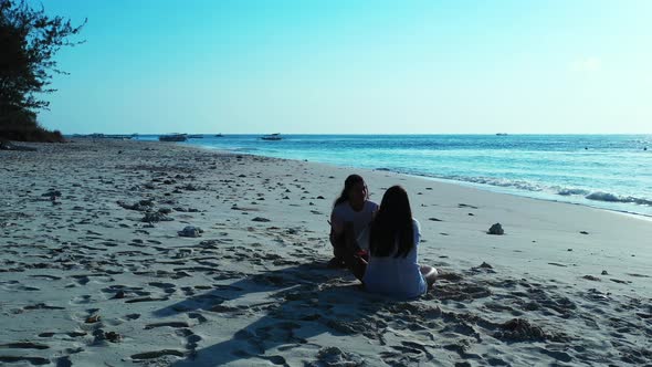 Girls in t-shirts sitting on white sandy beach, talking to each other about vacations on tropical is