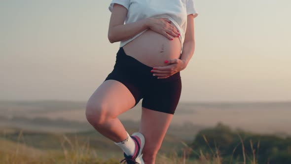 White Pregnant Woman in Sports Tshirt and Shorts Stands in Yoga Pose and Holds Her Stomach Closeup