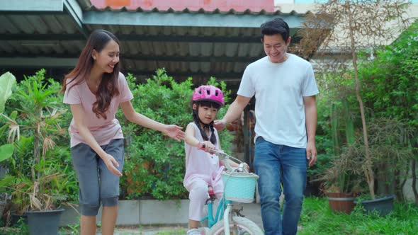 Asian couple father and mother teaching small pretty kid daughter in helmet riding on bike at street alt