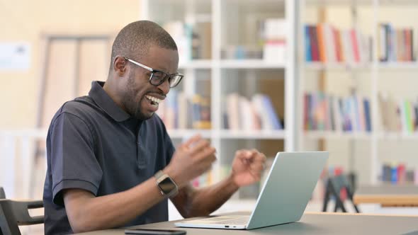 African Man Celebrating Success on Laptop in Library