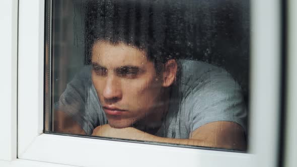 Sad beautiful young guy is at home behind the window during the rain. Depression and loneliness