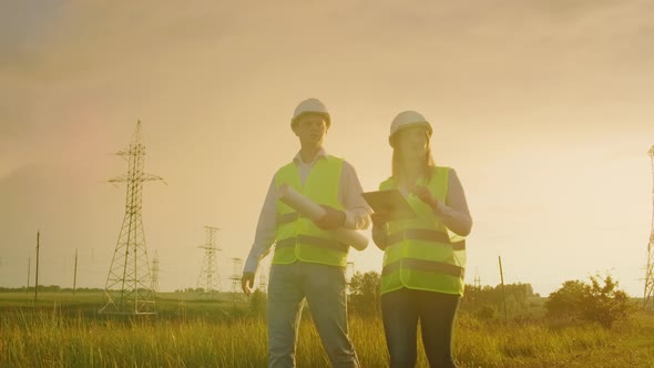 Two Electricians Work Together Standing in the Field Near Electricity Transmission Line in Helmets