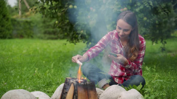 Young Woman Using a Smartphone While Sitting By a Campfire on a Picnic