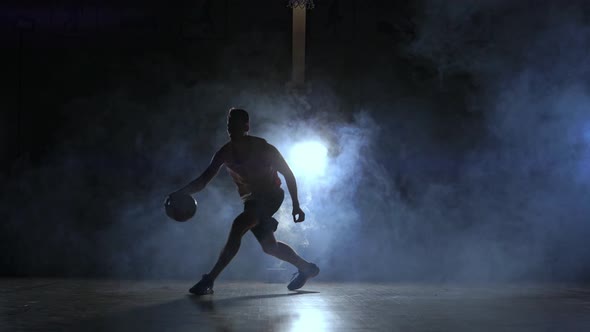 A Man with a Basketball on a Dark Basketball Court Against the Backdrop of a Basketball Ring in the