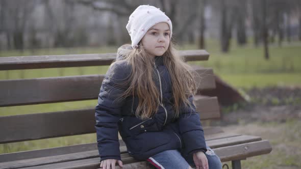 Brunette Schoolgirl Sitting on Bench in Park and Raising Leg. Portrait of Lonely Sportive Kid