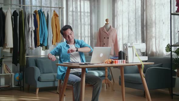 Asian Male Designer Working On A Laptop And Stretching While Designing Clothes In The Studio