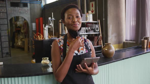 Portrait of african american woman in face mask working at a bar, using tablet and smiling to camera