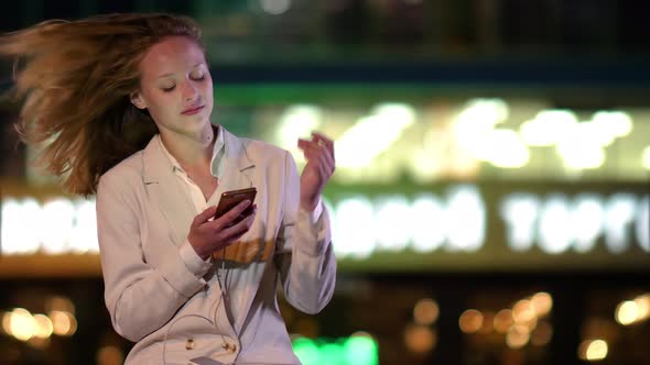 Beautiful Woman Using Her Phone Late at Night When Lights Are Turning Off in the Building Behind Her